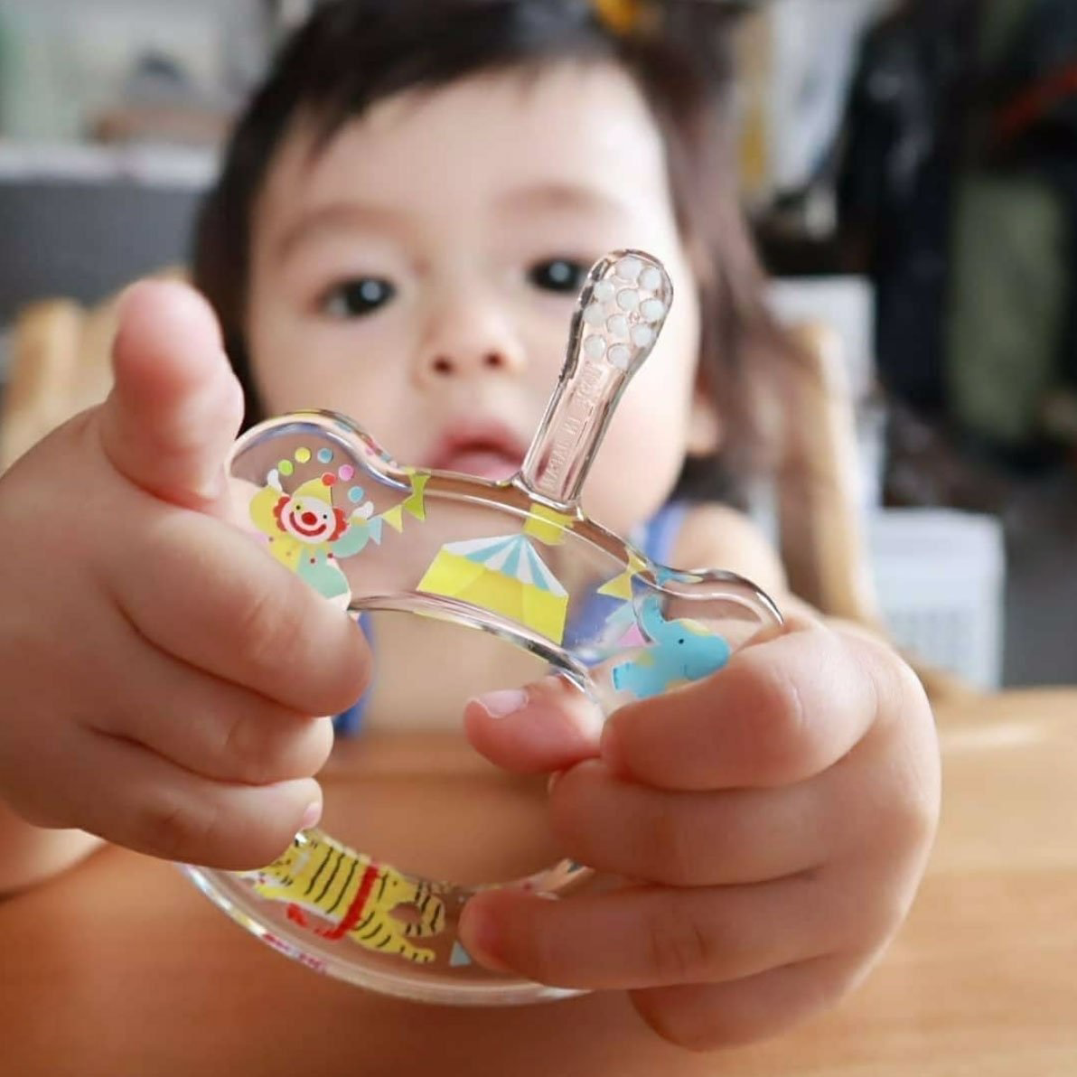 A young child sits at a table, holding a small, clear plastic toy adorned with colorful animal and circus-themed stickers. The child is intently focused on the toy, their face slightly out of focus in the background. Nearby lies one of the vibrant HAMICO - BABY TOOTHBRUSH UNICORNS by HAMICO/INNOVATIVE TOOTHBRUSH, designed with an ergonomic handle for easy grip.