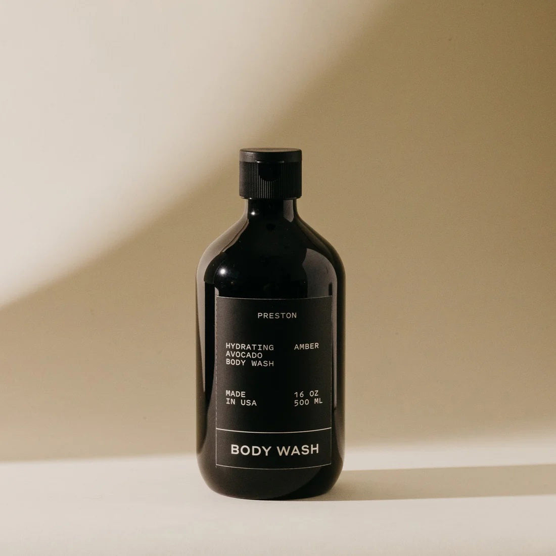 A black bottle of PRESTON - BODY WASH labeled "Hydrating Avocado Body Wash Leather" is centered against a beige background. Made in the USA with plant-based ingredients, this 16 oz (500 ml) moisturizing body wash from the brand PRESTON ensures hydrated skin without harsh chemicals.