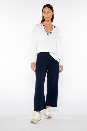 A person stands against a plain white background wearing the KINROSS CASHMERE GATHERED SLEEVE SPLITNECK SWEATER and navy blue wide-leg pants. Their long hair cascades down, and they sport white sneakers with beige soles, with hands resting relaxed by their sides.