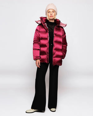 A person stands against a plain background, wearing the MIRTO 1956 - WATER REPELLENT QUILTED JACKET in a shiny red color, which features a removable hood. They are also dressed in black wide-leg pants and a light pink beanie. Their left hand hangs relaxed by their side while their right hand holds the jacket. Facing forward with a neutral expression, they complete the look effortlessly.