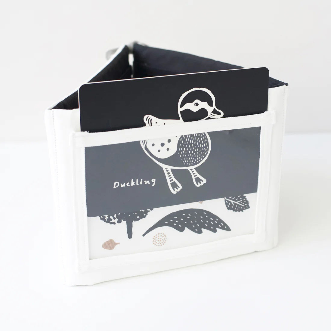 A TUMMY TIME GALLERY soft book from WEE GALLERY, featuring black and white fish and squid illustrations, is partially inserted in a clear front pocket. A brown tag attached by a string reads &quot;tummy time gallery.&quot; This Montessori toy, ideal for sensory development, rests on a plain white surface.