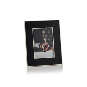 The BLACK BONE INLAY WITH WHITE INNER BORDER tabletop picture frame by ZODAX, measuring 5 in x 7 in, features a stand and displays a photo of a person sitting indoors on a bed. The frame has a white border around the slightly blurred image and is set on a white, reflective surface.