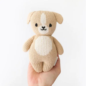 The CUDDLE + KIND - HANDMADE BABY PUPPY by CUDDLE & KIND is displayed from the back, highlighting its beige knitted body, small floppy ears, stubby arms, and short tail with no visible facial details. Meticulously hand-knit from 100% cotton yarn, its texture is distinctly visible, enhancing its cozy and handcrafted appeal. Perfect for those who appreciate fair trade and hand-knit baby animals.