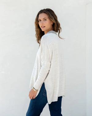 A person with wavy brown hair stands sideways against a plain white background, wearing a light-colored, loose-fitting women's sweater and blue jeans. They glance towards the camera with a neutral expression, showcasing the sporty and stylish appeal of the MERSEA - CATALINA POLO SWEATER by MER SEA.