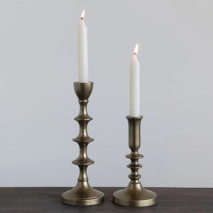 Two aluminum taper candle holders, both from Creative Coop, are shown against a white background. The candlestick on the left is taller and features an intricate, vintage design with multiple curves, identified as the Aluminum Taper Candle Holder (Large), while the shorter one on the right boasts a simpler style reminiscent of an antique gold candle holder.