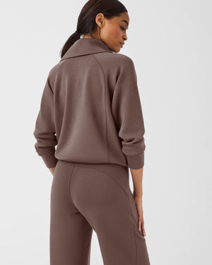 A woman with long brown hair is wearing the SPANX AIRESSENTIALS HALF ZIP sweatshirt and matching pants, both crafted from lightweight fabric for luxurious comfort. She holds her braided ponytail with one hand while looking to her right against a plain white background.