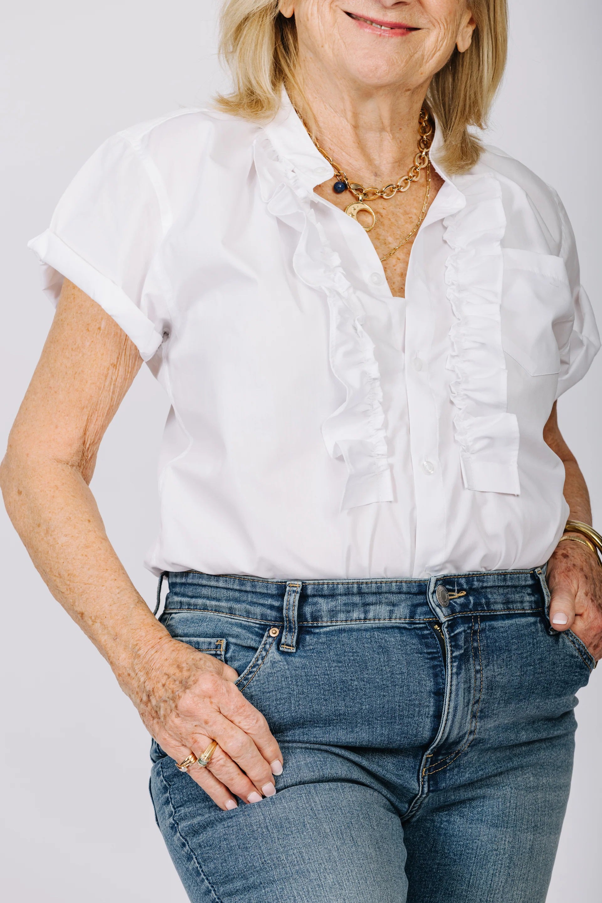 A person wearing the KMJ - KIKI SHIRT, a white ruffled blouse, and blue jeans stands with hands on hips, embodying a refined style. They accessorize with gold bracelets, rings, and a necklace. Their nails are neatly manicured and painted white. The background is plain.