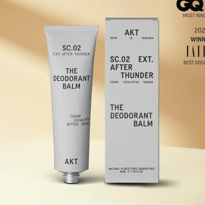 A tube and box of AKT LONDON AKT DEODORANT BALM AFTER THUNDER, featuring cedar, eucalyptus, and orange, provide all-day protection. The minimalist packaging highlights its aluminum-free formula on a beige background.