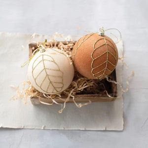 Nestled in a wooden box with straw-like filling on a light-colored surface are two VELVET BALL ORNAMENT 3" by CREATIVE COOP, one in white and the other in copper, both adorned with glittering gold leaf patterns. These ornaments add an elegant touch to any holiday decor.