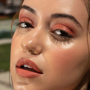 A close-up of a person with curly dark hair, a serious expression, and warm makeup featuring the universally flattering, weightless JASON WU - HOT FLUFF LIP/EYE/CHEEK by JASON WU BEAUTY on their face. They are looking directly at the camera with a neutral background.