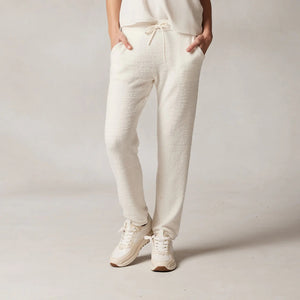 A person stands against a plain background, wearing a white sleeveless top and KASHWERE's RELAXED JOGGER in an ivory color. The joggers are made from lightweight, breathable Kash-ease yarn and have pockets where they’ve placed their hands. The individual completes the look with white sneakers.