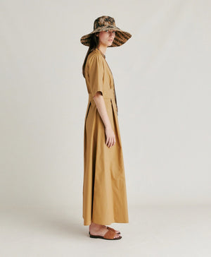 A woman stands in profile wearing the MOMONI - MARTINELLA DRESS, a long, tan cotton poplin dress from MOMONI with short sleeves and a flared skirt. She pairs it with a wide-brimmed, patterned hat, brown sandals, and a necklace. She faces left against a plain, light background.