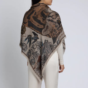 A person draped in the SABINA SAVAGE - THE LUNAR LEOPARD CASHMERE SHAWL 135CM by SABINA SAVAGE ENGLAND, showcasing an animal print design reminiscent of samurai armour. The shawl is adorned with a prominent image of a leopard complemented by illustrations of birds and various patterns. The individual is styled in a beige top and white pants, standing against a plain background.