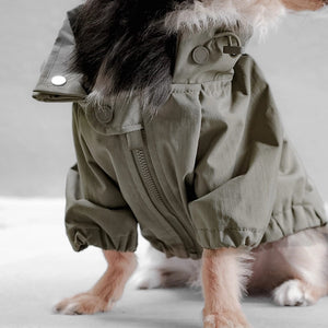 A small dog is wearing an oversized, olive-green, water-resistant jacket with a fur-lined collar. The dog's legs and part of its face are visible, while the rest of its body is covered by the LAMBWOLF COLLECTIVE - BROOKLYN LIGHT & PACKABLE DOG JACKET. The background is a soft, neutral color.