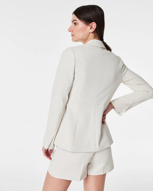 A woman wearing the SPANX - CAREFREE CREPE WRAP BLAZER, paired with a white top. She holds the lapel to reveal the inner lining of the tailored crepe blazer. With a neutral expression and her hair in a low ponytail, she stands against a plain white background.