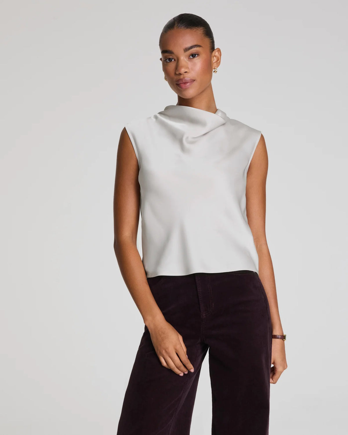 A woman wears the SPANX Silky Smooth Cowlneck Sleeveless Top in light gray with dark high-waisted pants, sitting on a stool against a plain background and looking at the camera with a neutral expression.