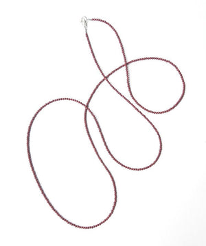 The ANN LIGHTFOOT - RED JASPER STRAND NECKLACE by ANN LIGHTFOOT, featuring small, round dark red beads, is laid out on a white surface with its 39-inch length forming loose curves. This delicate piece is fastened with a sterling silver clasp.