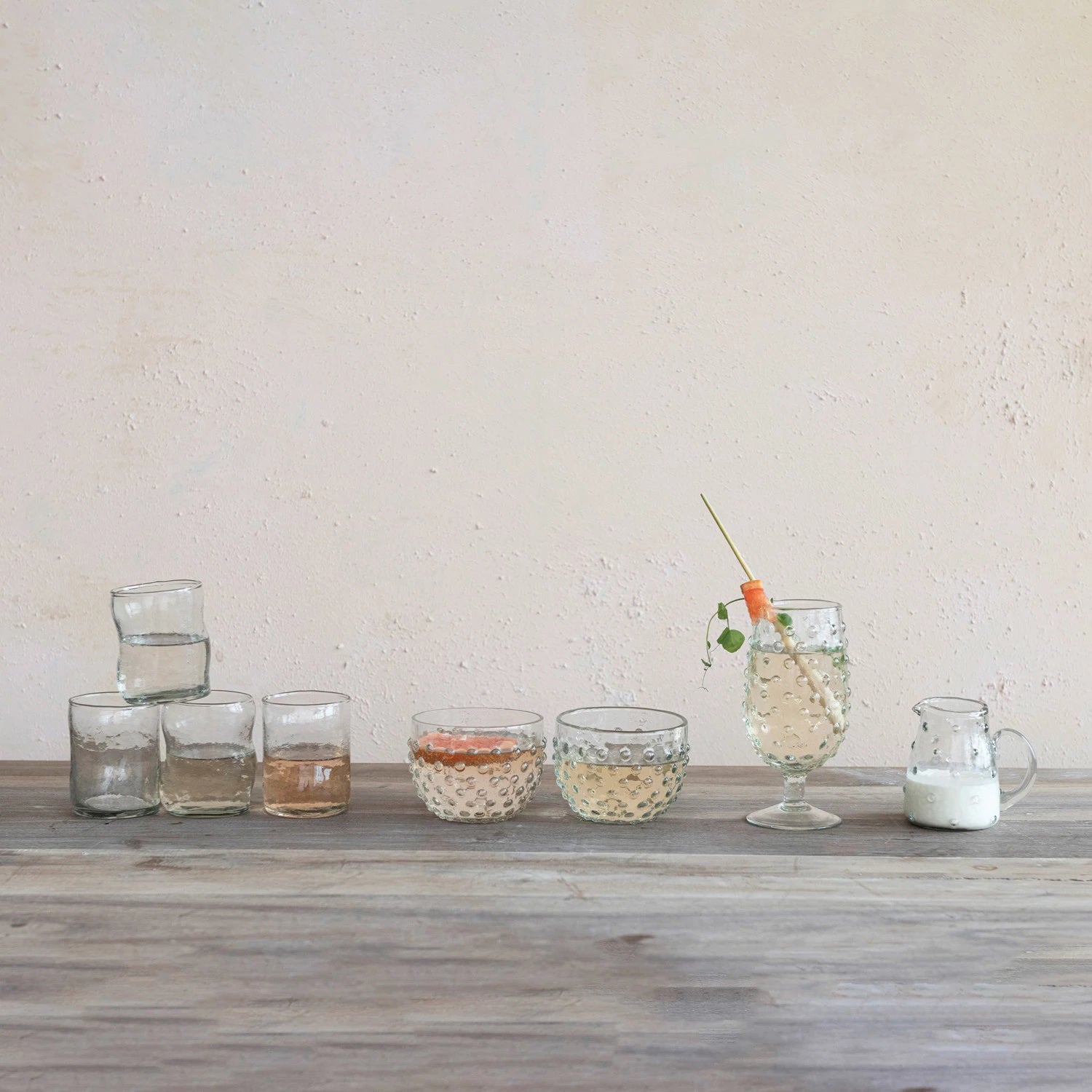 A variety of Creative Coop's textured glassware rests on wood: stacked small glasses with liquid, a single glass, two glasses with different liquids, a large glass with garnish, and an eco-friendly Recycled Glass Hobnail Creamer filled with milk against a neutral wall.