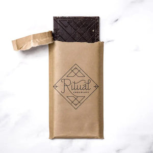 The RITUAL CHOCOLATE - SMORES CHOCOLATE BAR 70% features a rectangular shape adorned with a speckled golden topping, reminiscent of marshmallow flavors, set against a white marble background. The bar has a slightly textured surface due to the glittery gold flecks covering it.