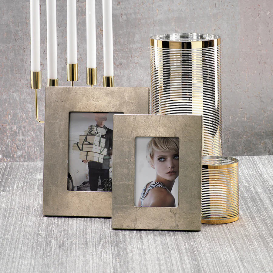 Two ZODAX SILVER LEAF PHOTO FRAMES - 5x7, each displaying black-and-white images, rest on a textured surface. Behind them, tall candle holders with silver-and-gold stripes and a metal candelabra holding lit taper candles form an elegant arrangement against the gray, textured wall.