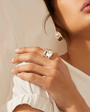 The JENNY BIRD - VIVIANA RING is a versatile accessory flaunting a wide silver band and an elegant, smooth, wavy design. The inner side of the ring is engraved with "JENNYVI," while the backdrop remains plain and light-colored.