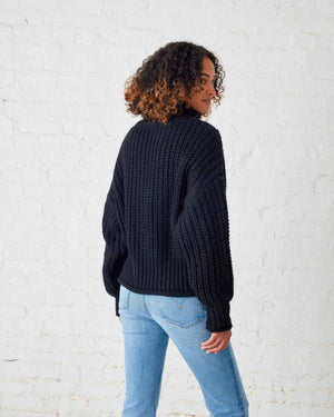 A woman with curly hair stands against a white wall, wearing a chunky mustard-colored MERSEA - BEACHSIDE TURTLENECK and blue jeans. She holds the hem of her turtleneck with one hand and has a relaxed expression.