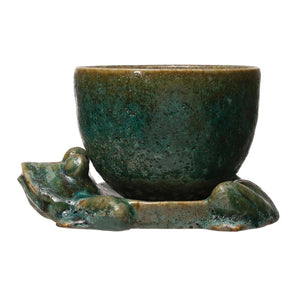 The CREATIVE COOP STONEWARE PLANTER features a green ceramic cup with textured surface, resting on a lounging frog-shaped base. The reactive glaze highlights the frog's limbs, which form part of the support, adding organic charm.