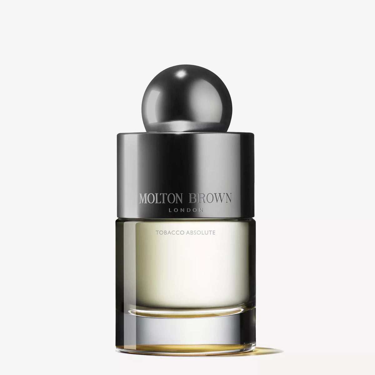 A clear glass bottle of MOLTON BROWN - ABSOLUTE TOBACCO EAU DE TOILETTE 100ML stands upright with a metallic silver cap and rounded top, set against a plain white background, reflecting the refined essence of this tobacco perfume.