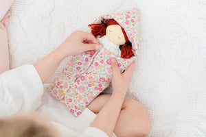 A child wearing a white long-sleeved shirt is seen holding the ALIMROSE - MINI MATILDA ASLEEP AWAKE doll, which has red hair and is snugly tucked into a floral-patterned sleeping bag. The cloth doll features a hood covering its head. The background displays a white textured surface, encouraging developmental play and social skills.