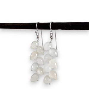 A pair of delicate ANN LIGHTFOOT - LEAF PETAL WHITE MOONSTONE EARRINGS is hanging from a dark rod. Each earring features multiple small, white moonstone teardrop-shaped beads arranged in a cascading design, attached to polished sterling silver earwires. The background is plain white, highlighting the earrings' elegance.