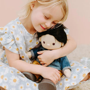 A Dinkum Doll from Olli Ella USA's Travel Togs collection, featuring dark hair and dressed in a denim jacket, mustard pants, and black shoes, stands on a wooden surface. Beside the doll is a teal See-Ya Suitcase with white trim and hardware. In the background, another larger teal suitcase is partially visible.