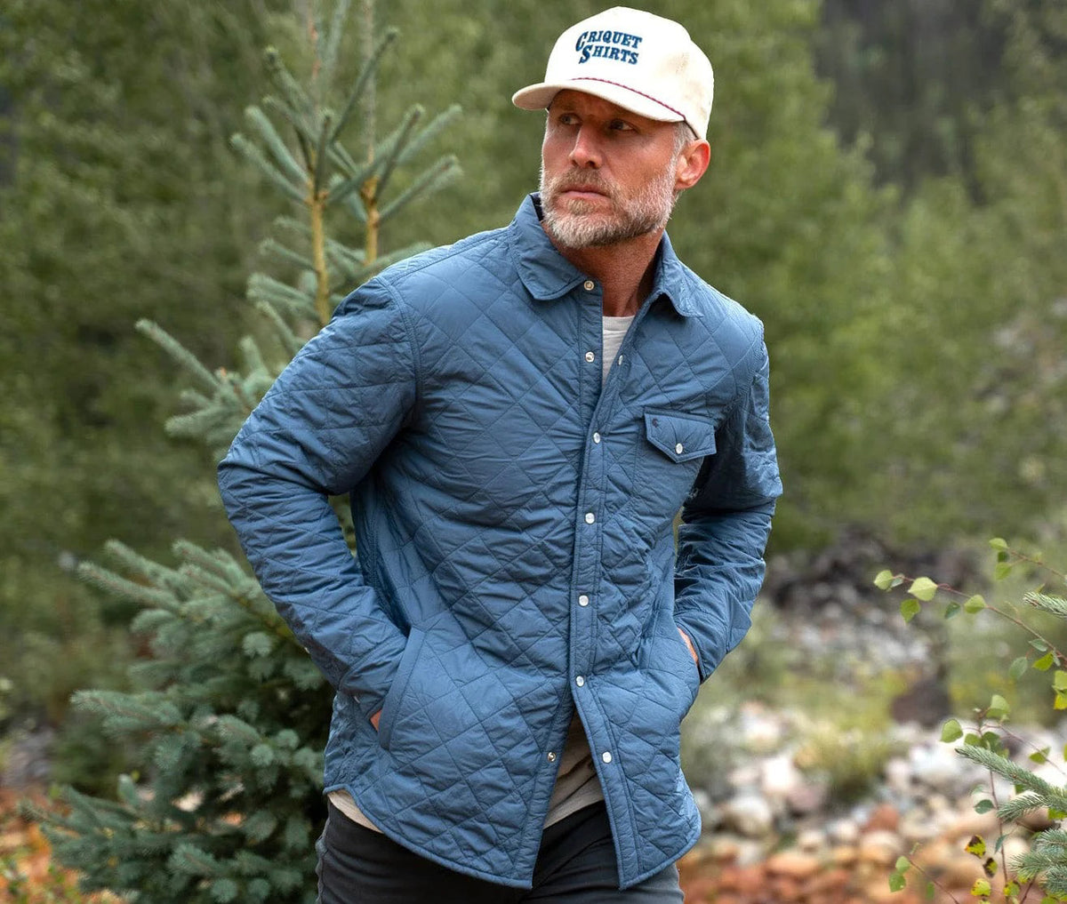 A man with a gray beard wears the CRIQUET - QUILTED SHACKET, a light blue piece with a pearl snap closure, along with a cream cap. He stands outdoors among pine trees and greenery in a forested area.