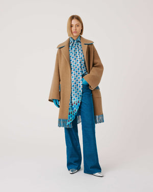 A person is wearing the SFIZIO - TWO TONED COAT WITH FRINGE by SFIZIO, featuring blue and brown fringe patch pockets and hem. They are also donning blue gloves and blue pants. The image is cropped to show from the shoulders down against a plain, off-white background.