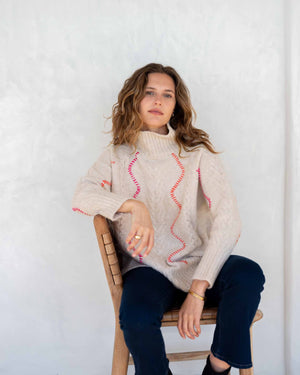 A person with long hair is wearing the MERSEA - ALPINE SWEATER by MER SEA, a loose-fitting, light grey acrylic sweater adorned with subtle purple zigzag patterns. They are also dressed in light blue jeans. The background is plain white.