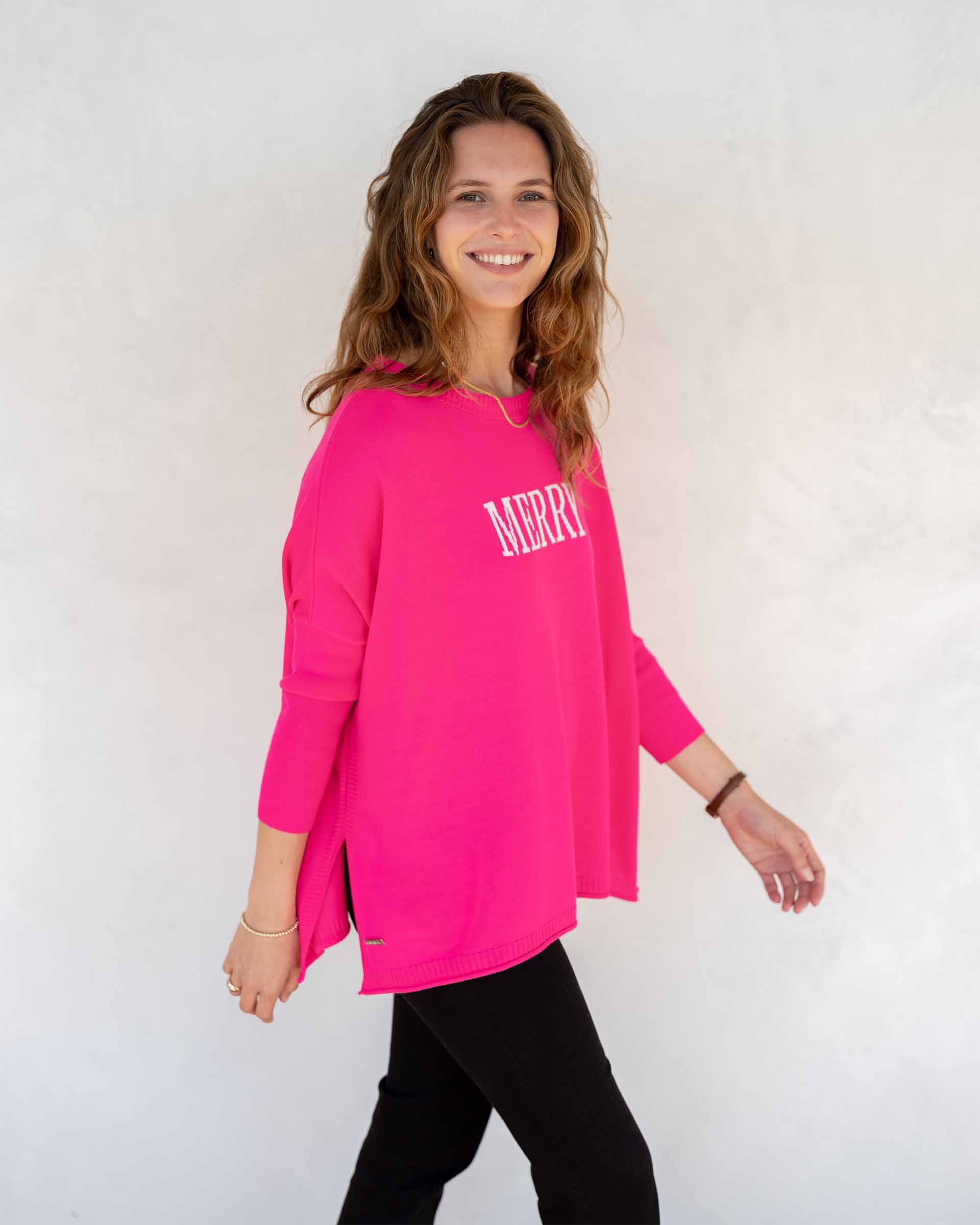 A person with long brown hair is wearing the stylish CATALINA CREWNECK SWEATER MERRY from MER SEA, featuring "MERRY" in white on a vibrant pink fabric, capturing the essence of a true MERSEA staple. The look is elegantly finished with a thin gold necklace against a simple, light-colored backdrop.