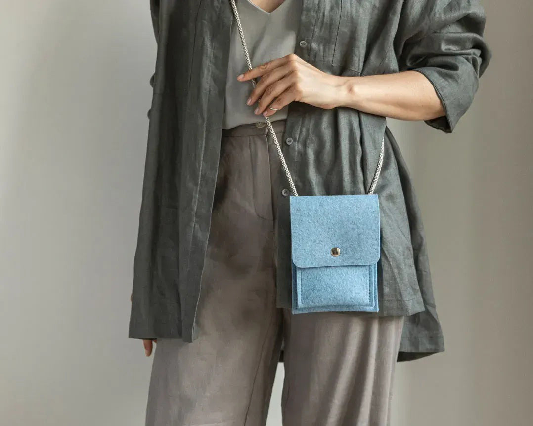 A person in a loose gray shirt and taupe pants carries the WOOLBERRY WOOL FELT PHONE BAG—a pink crossbody with a flap and silver chain—over their shoulder, showing only their torso and part of their arms.