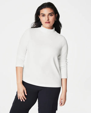 A person with long, dark hair smiles and poses with one hand in their pocket. They are wearing the SPANX - AIRESSENTIALS MOCK NECK TOP made from super-soft fabric, paired with matching dark green leggings against a light background.