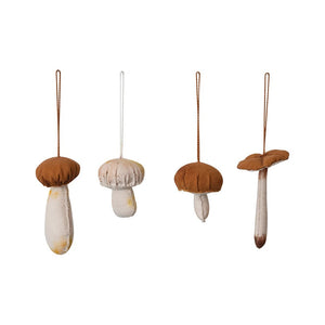 Amidst a textured backdrop sits a square wooden box brimming with small round seeds. Nestled within are five HANDMADE COTTON MUSHROOM ORNAMENTS by CREATIVE COOP, each featuring brown caps and light stems that highlight their exquisite stitching craftsmanship. Some of these mushrooms are adorned with strings, enhancing the display of four distinct styles.