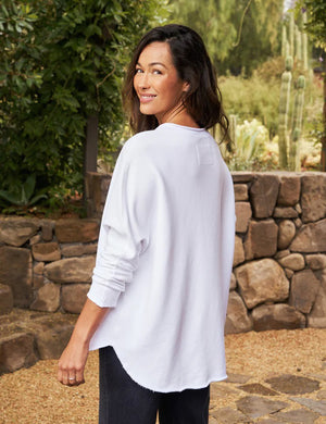 A person with long, wavy blonde hair is standing outdoors near a large tree, wearing the FRANK & EILEEN ANNA LONG-SLEEVE CAPELET in navy over a white shirt and blue pants. The background showcases green foliage and a stone path. The person is smiling and gazing to the side.