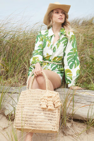 A woman in a green and white leaf-patterned outfit sits on a wooden log by the beach, wearing a straw hat. She holds the MAR Y SOL - GEORGIA TOTE adorned with raffia tassels on her lap. Tall grass and sand can be seen in the background.