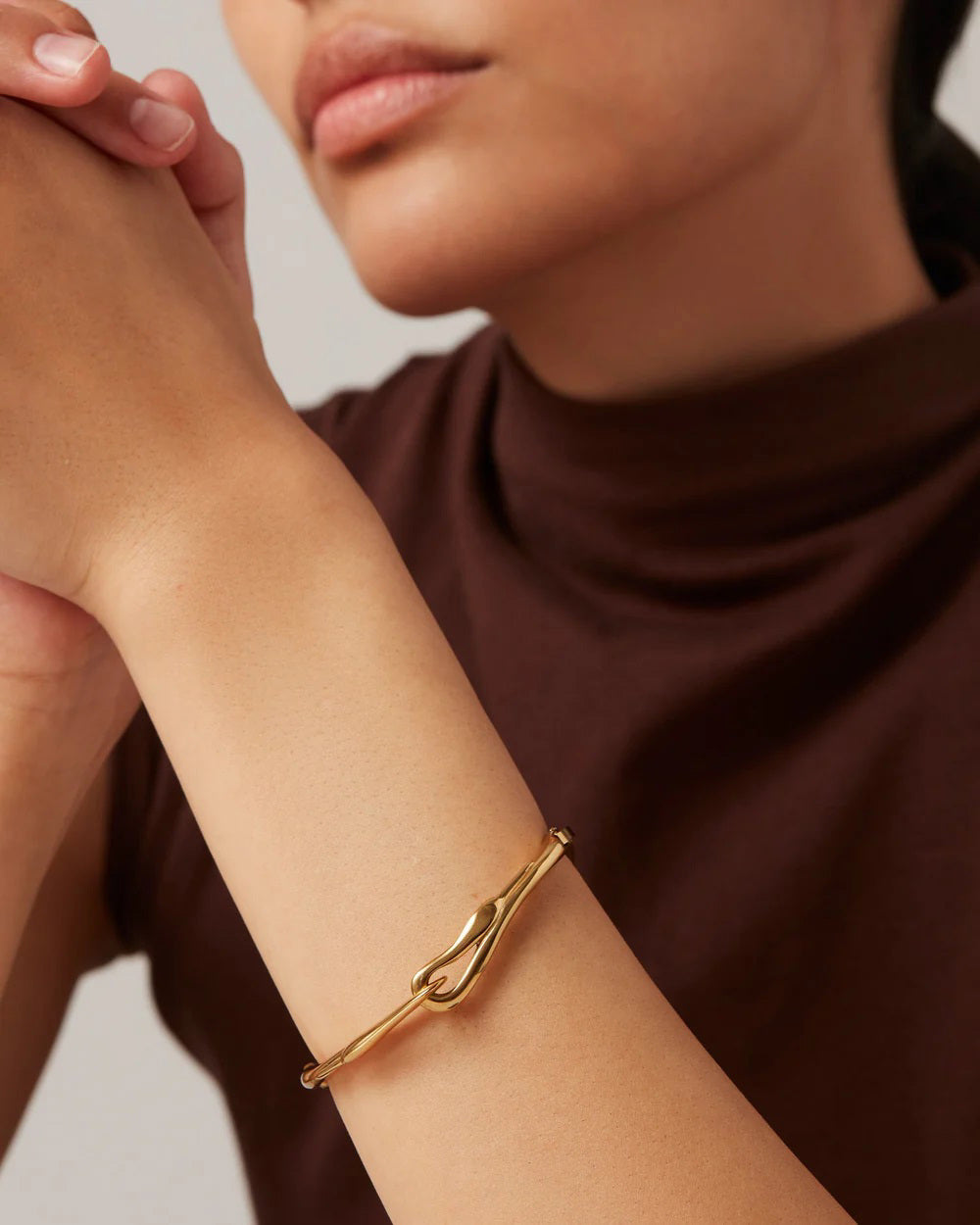 Wearing the JENNY BIRD Lisieux Bangle, designed like a horse bit with an elegant foldover clasp, a person rests their chin on clasped hands. They're dressed in a brown turtleneck sweater against a neutral background.