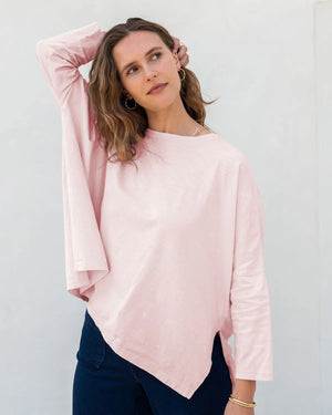 A person with long, wavy hair is smiling and posing with one hand touching their face. They are wearing the oversized fit, long-sleeved black MERSEA - CATALINA SLUB TEE and matching black pants made from a lightweight cotton/polyester blend. The background is plain white.