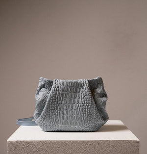 A black, textured handbag with a rectangular base and a gathered top is displayed on a white pedestal. The handbag, designed by DANIELA LEHAVI and named the TOKYO BALI BAG, features an alligator-like pattern and appears to be made of leather or a similar material. The background is plain and neutral-colored.