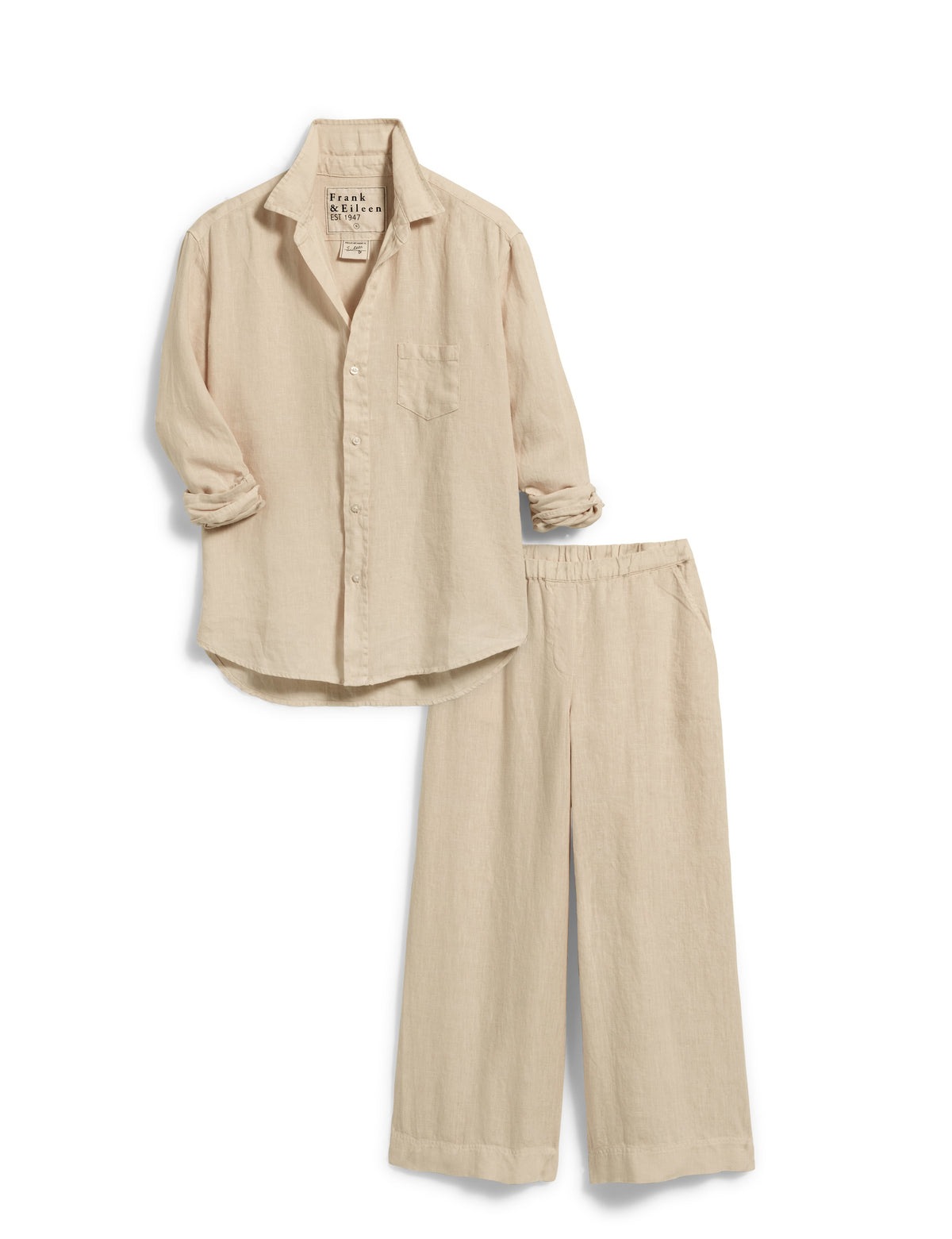 A woman with curly hair, wearing sunglasses, a beige long-sleeve shirt, and a matching pair of FRANK &amp; EILEEN Maisie Wide Leg Linen Pull On Pants from the Tuscany Getaway Set, stands outdoors. She is smiling and leaning against a rustic stone wall. Nearby is a table with a wine bottle and glass, and a white chair under a lush green tree.