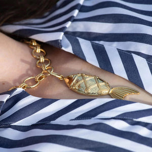 A person wearing the CAPUCINE DE WULF - POISSON PENDANT, featuring large interlocking rings of 18K gold over brass and a golden fish pendant, along with a matching gold ring. The person is wearing a blue top and their hand is touching the good luck talisman.