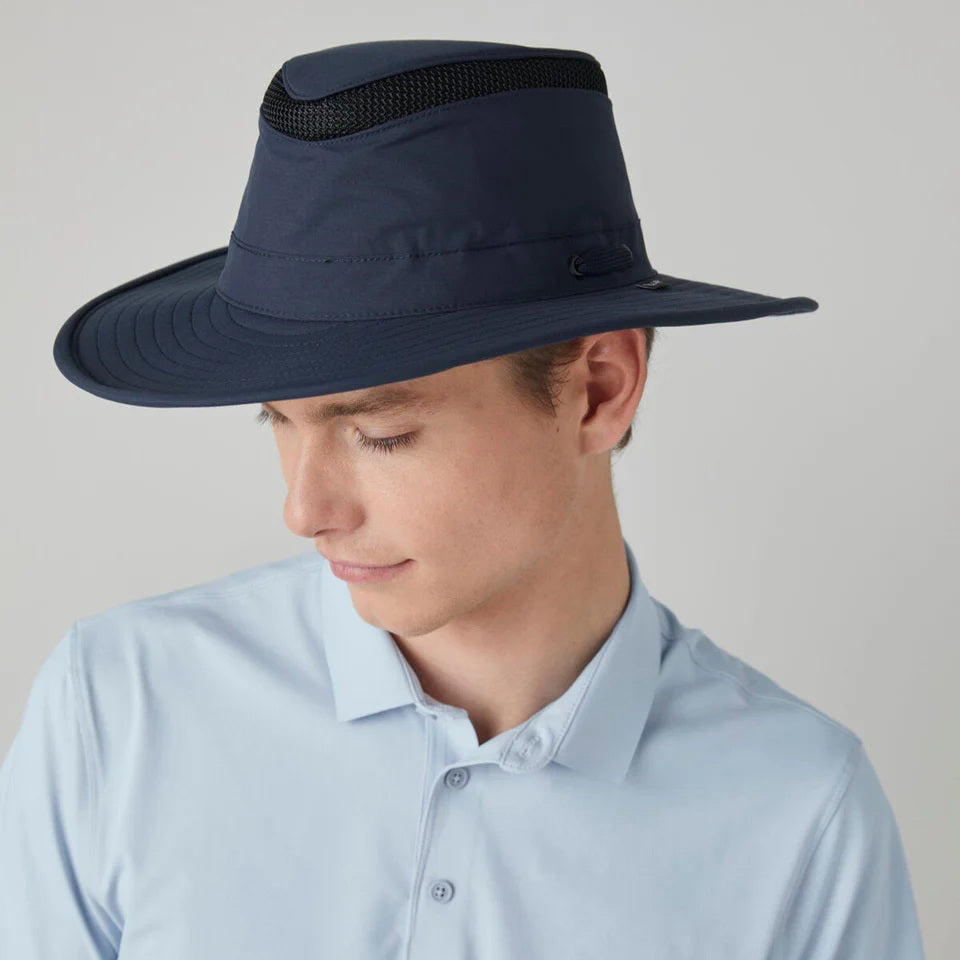 A person wearing the TILLEY - AIRFLO BROAD BRIM LTM6 hat with a light blue collared shirt, looking downward against a plain, light background.