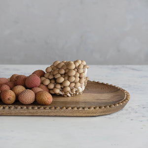 A MANGO TRAY W WOOD BEADS by CREATIVE COOP, crafted from 100% Mango Wood, holds a cluster of mushrooms and a pile of lychees, placed on a marble surface. The background is a light grey wall, creating a minimalistic and neatly presented arrangement.