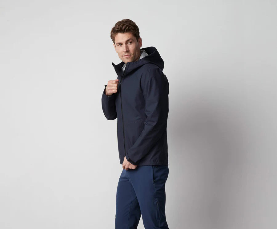 A man stands in front of a white wall wearing the MACK WELDON STORMCHASER JACKET with adjustable hood, paired with a gray shirt, blue pants, and black sneakers. He looks to the side while holding the jacket&#39;s zipper.