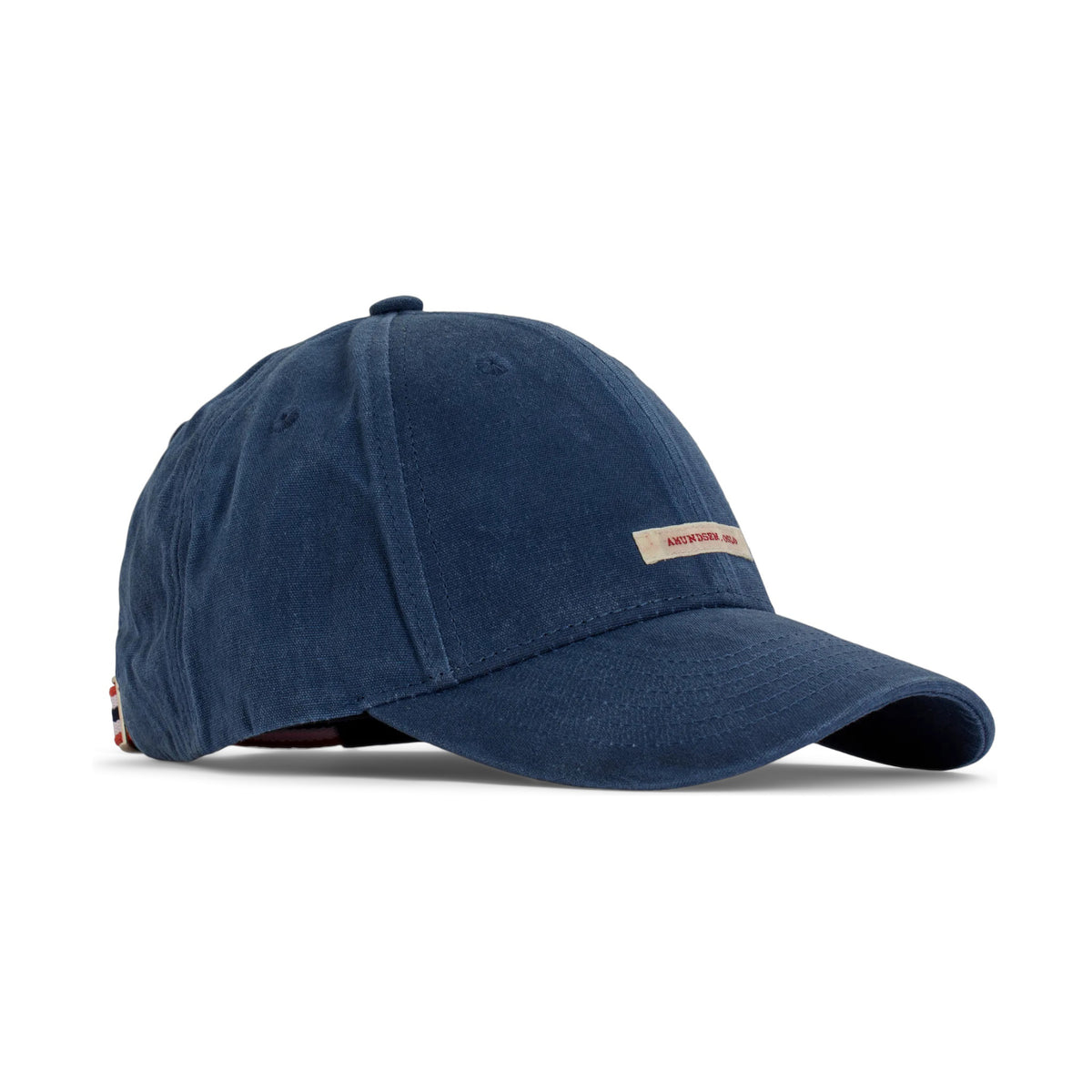 The AMUNDSEN SPORTS - WAXED COTTON CAP in navy blue is shown from the side against a plain white background, featuring a curved brim and an adjustable strap with a red, white, and black checkered norgesreima design.