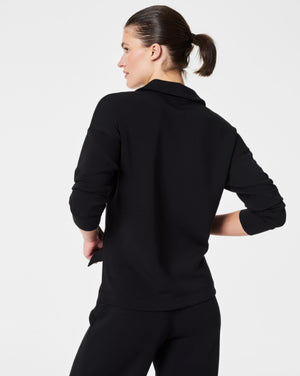 A person with dark hair styled in a neat updo is wearing the SPANX - AirEssentials Split Neck Polo Top in black, paired with matching black pants. They rest one hand on the back of their neck and gaze to the side against a plain white background, exuding an air of luxurious comfort.